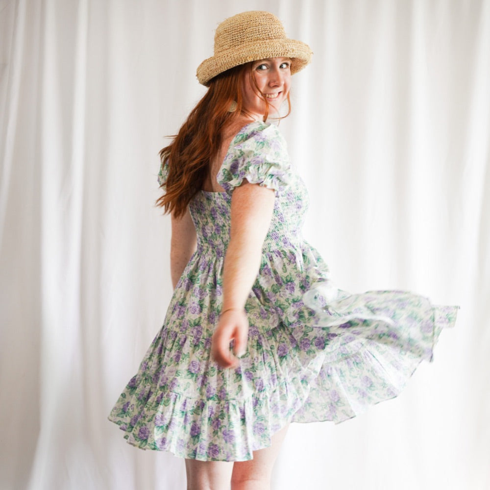 A woman wearing a purple and green floral print ruffle tier babydoll dress with short sleeves and a smocked bust stands in front of a white background. The dress is flowy and falls just above her knees. She is smiling and a raffia hat . The spring dress i