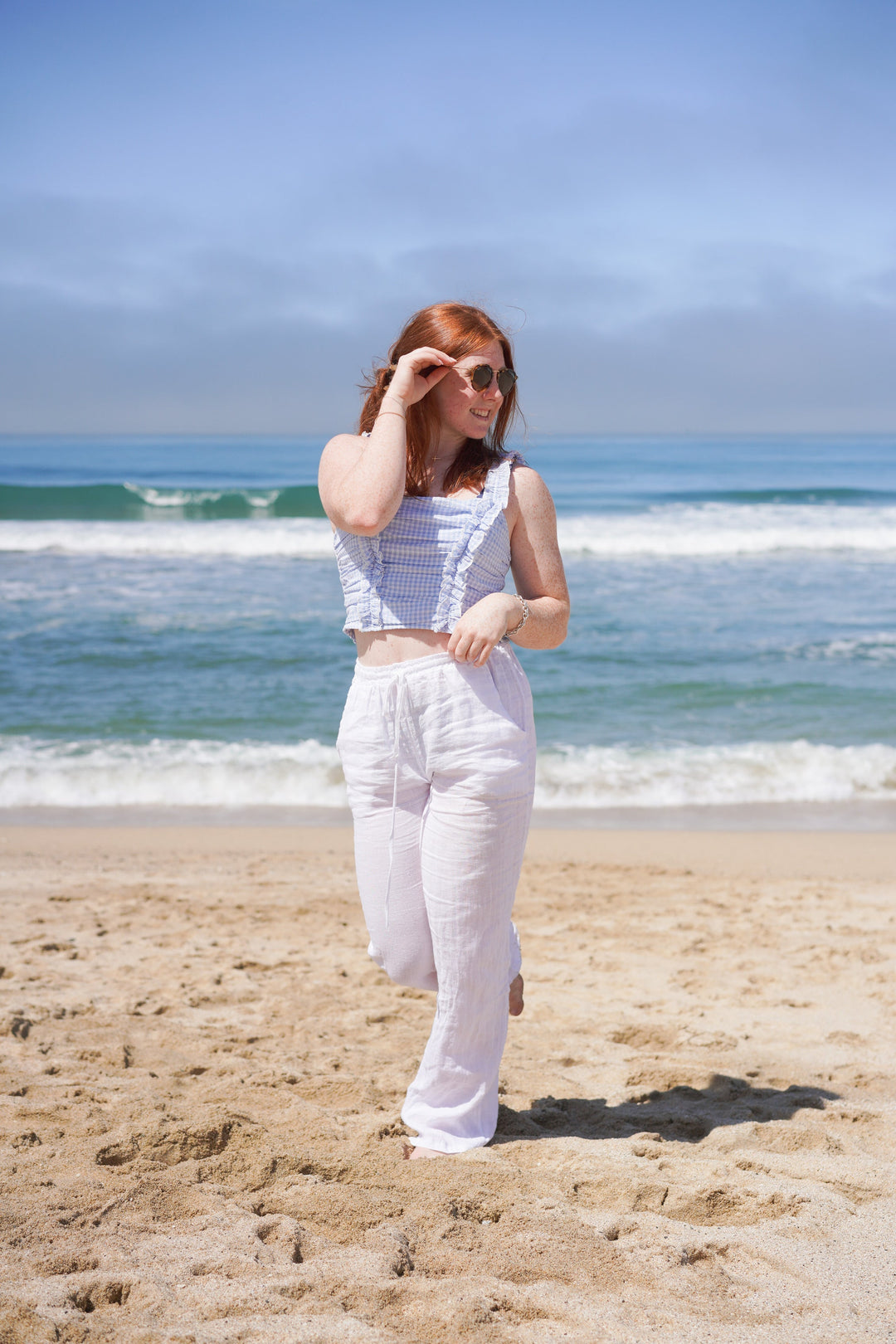 Girl walking on the beach wearing white linen pants.  Pants have a drawstring tie.  Top is cropped blue and white gingham