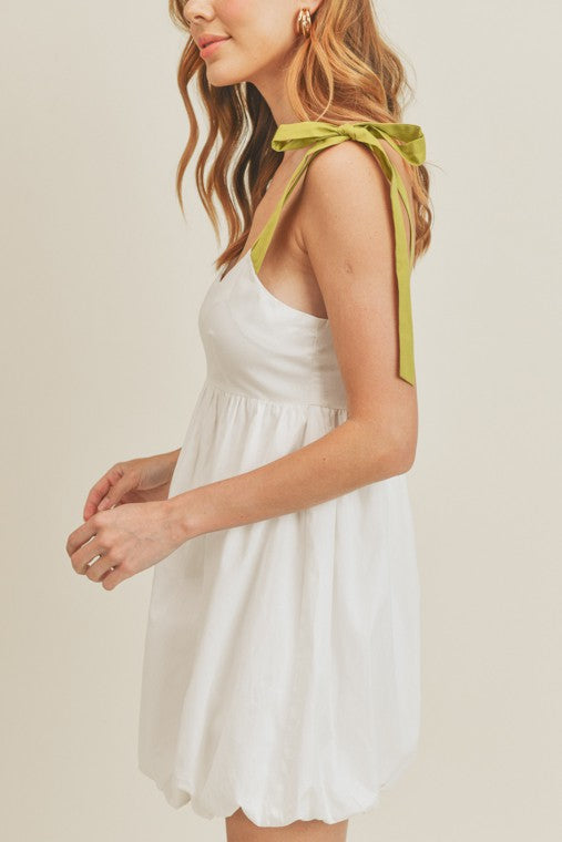 The model is wearing a white dress that has a light and airy feel to match the dress style. The dress is a bubble style with a full skirt and bubble hem. It has a pop of color and contrast to the body of it with it’s stunning green straps.