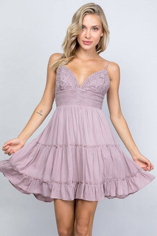 Model is wearing an ashy lilac color dress. The dress has a crochet lace bralette style top with adjustable spaghetti straps and an open back that ties in a bow. The skirt part of the dress is a light material that is tiered for a ruffle look.