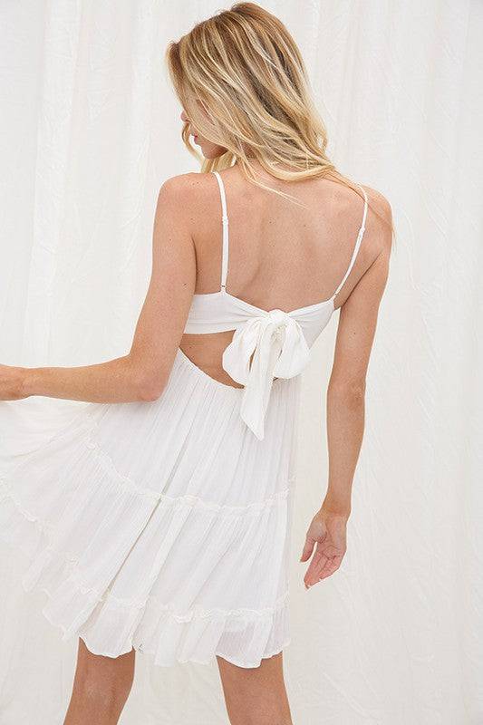 Model is wearing a ivory color summer dress. The dress has a crochet lace bralette style top with adjustable spaghetti straps and an open back that ties in a bow. The skirt part of the dress is a light material that is tiered for a ruffle look.