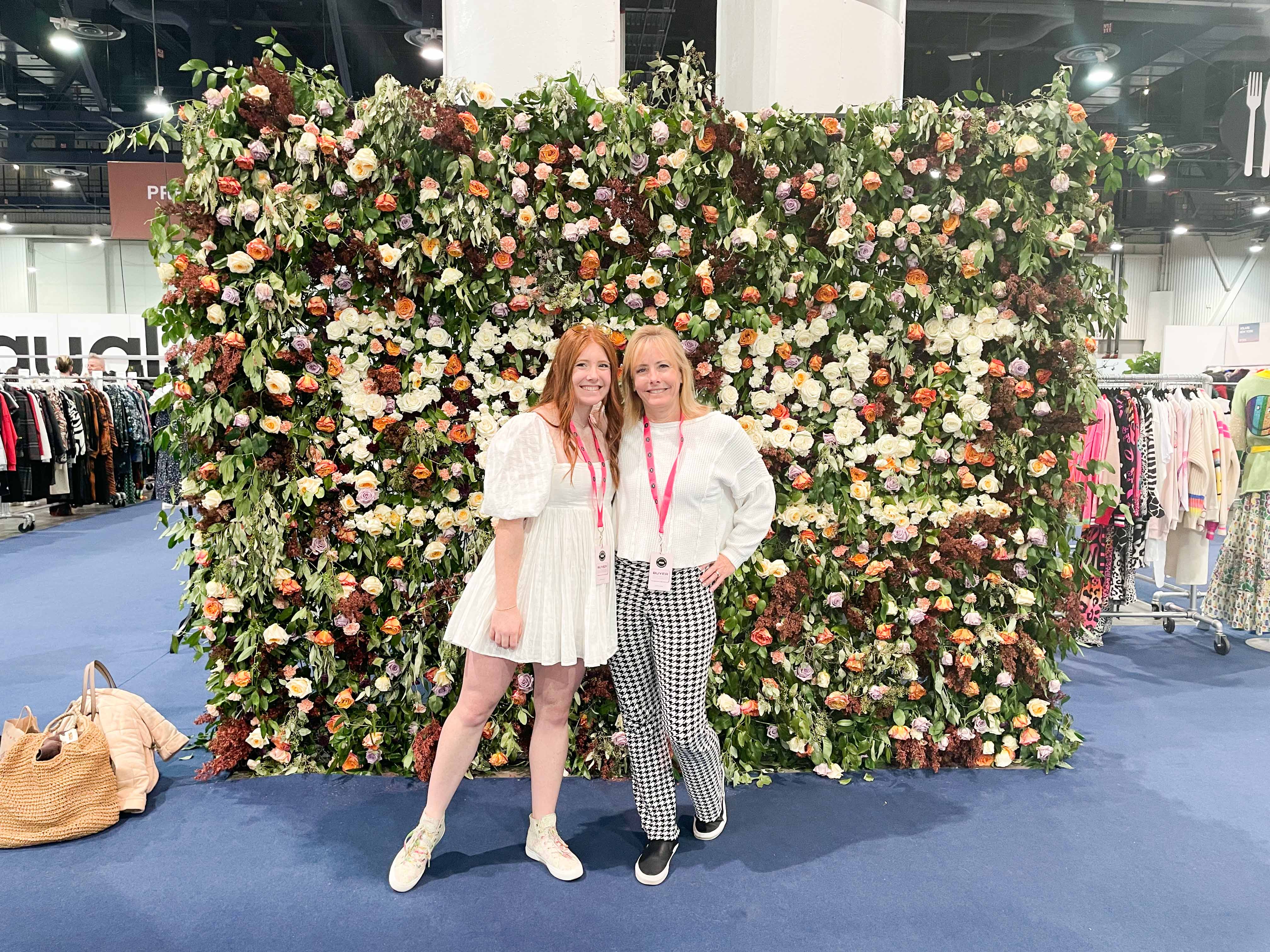 Two women standing in front of a beautiful flower wall at a convention. The woman on the left is wearing a white babydoll dress, while the woman on the right is dressed in houndstooth knit pants and a basic white long sleeve top. They are both smiling and appear to be enjoying the event.